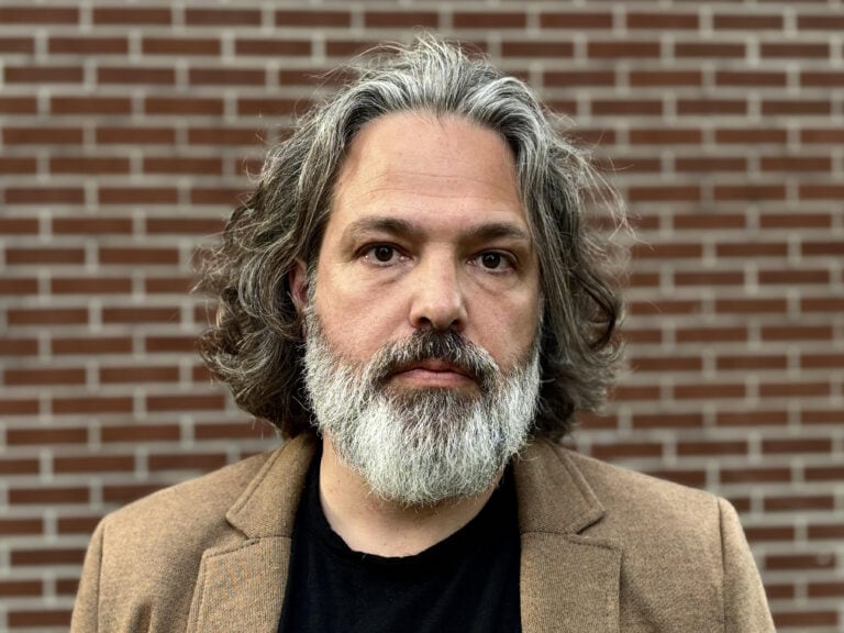 Thoughtful man with grey hair and beard, dressed in a brown blazer, standing in front of a brick wall, representing professionalism and style.